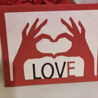 Close-up of handmade love card with red heart hands cut-out design and bold LOVE lettering on a white background.