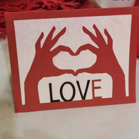 Handmade love card in red and white with heart hands design and the word LOVE on the front, displayed against a background of red hearts.