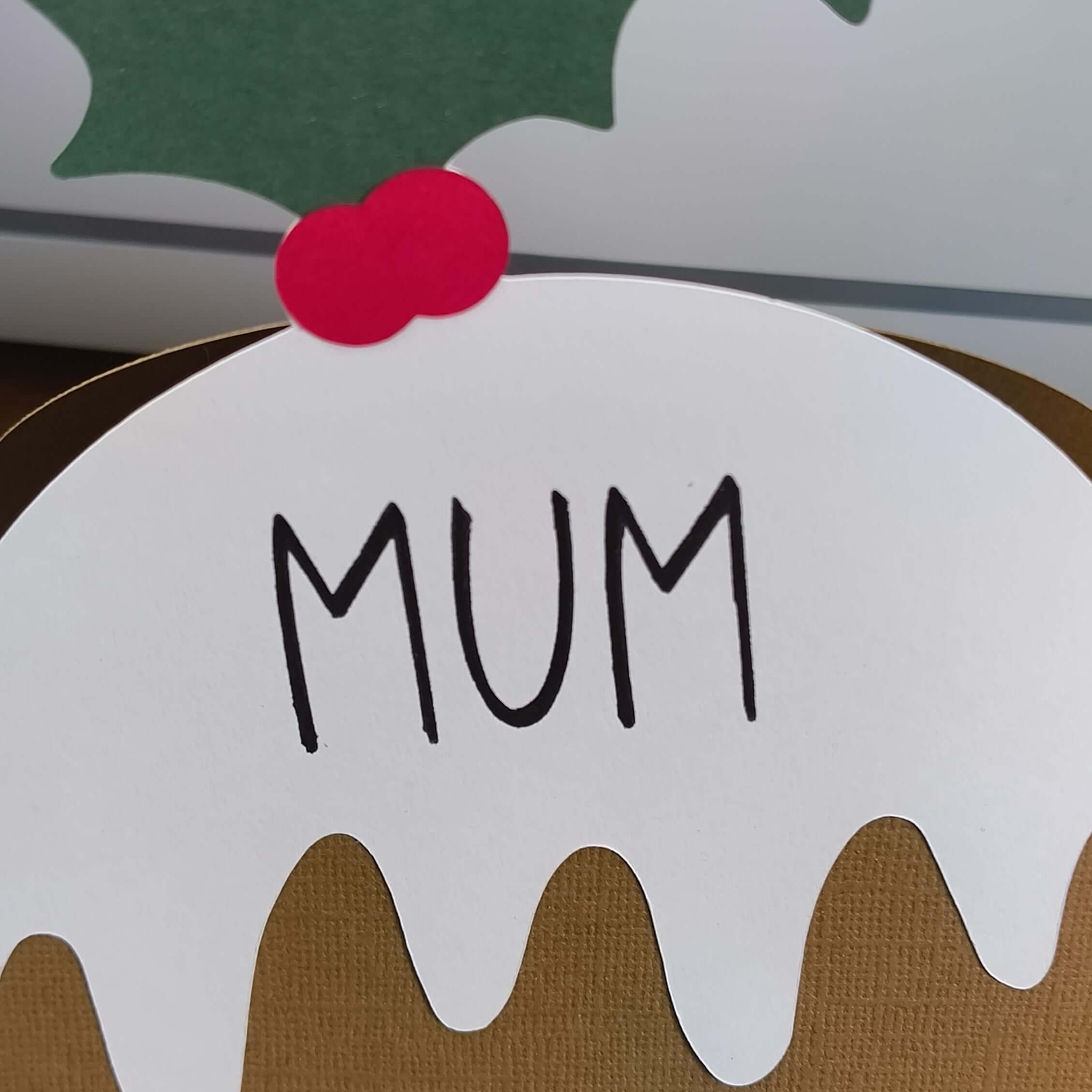 Handmade Christmas pudding place card featuring “Mum” text, with holly and berry topper, cut from layered textured cardstock in brown, white, green, and red.
