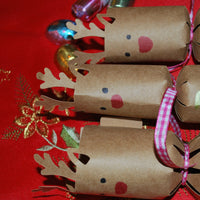 Group of handmade reindeer crackers with antlers, red noses, and pink gingham ribbons, personalised with names in gold foil, styled on festive red table linen.