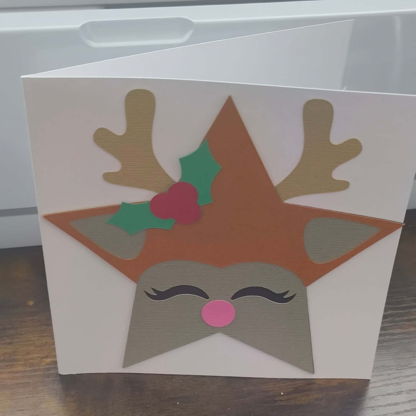 Handmade Christmas card featuring a reindeer with antlers, holly, and a pink nose on a star-shaped design.