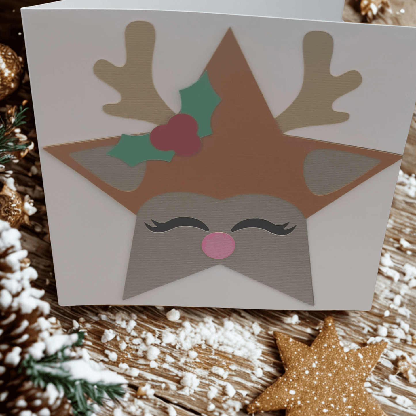 Handmade reindeer Christmas card featuring a star design, holly, antlers, and a pink nose on a festive background.