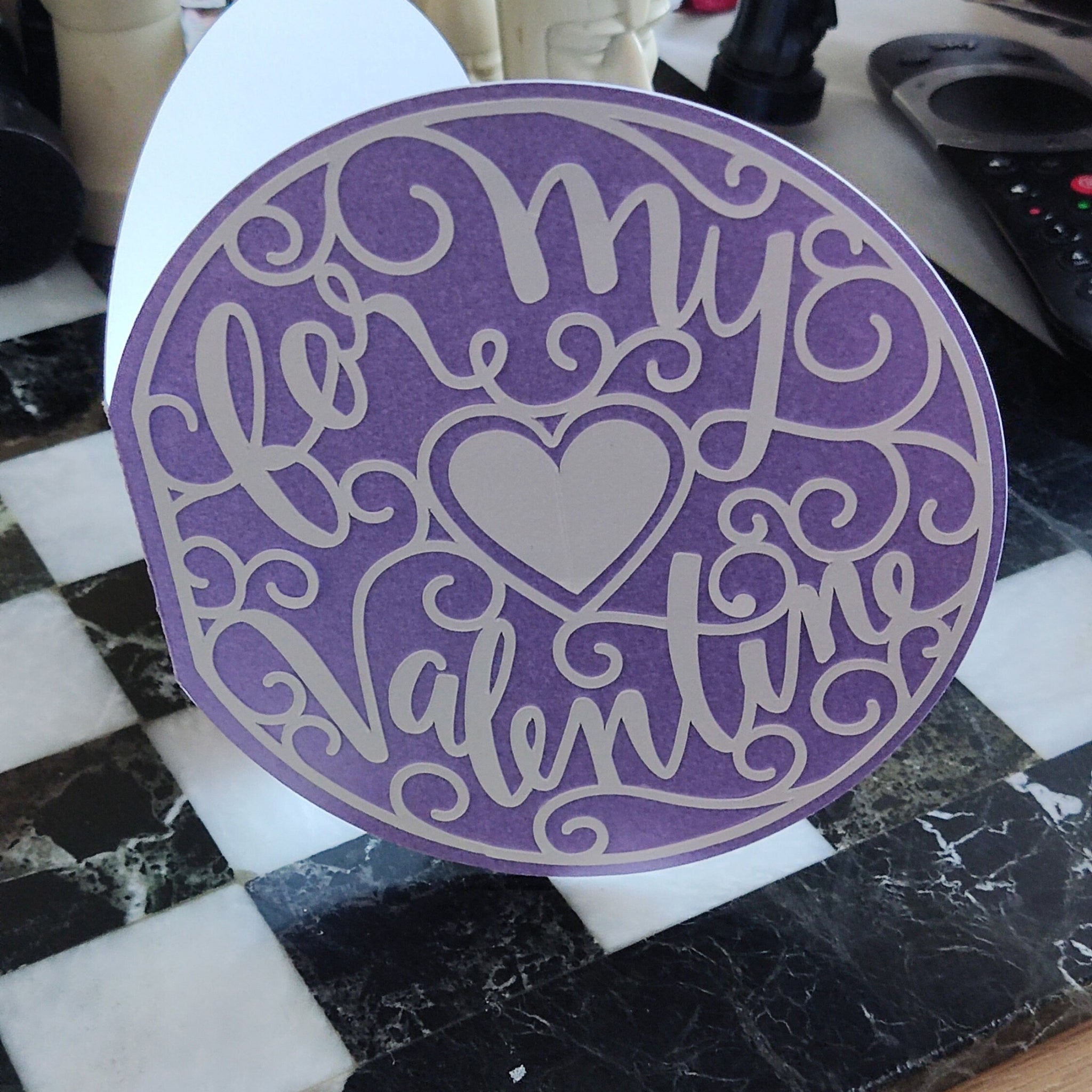 Circular purple Valentine’s card with white layered cut-out text and heart centrepiece, photographed upright on a table for full design visibility.