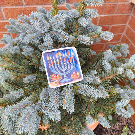 Hanukkah coaster with menorah and candles, displayed on a blue spruce tree outside against a brick wall