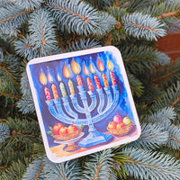Hanukkah coaster with menorah and fruit bowls, placed on a potted blue spruce by a brick wall