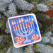 Hanukkah coaster with menorah and fruit bowls, placed on a potted blue spruce by a brick wall