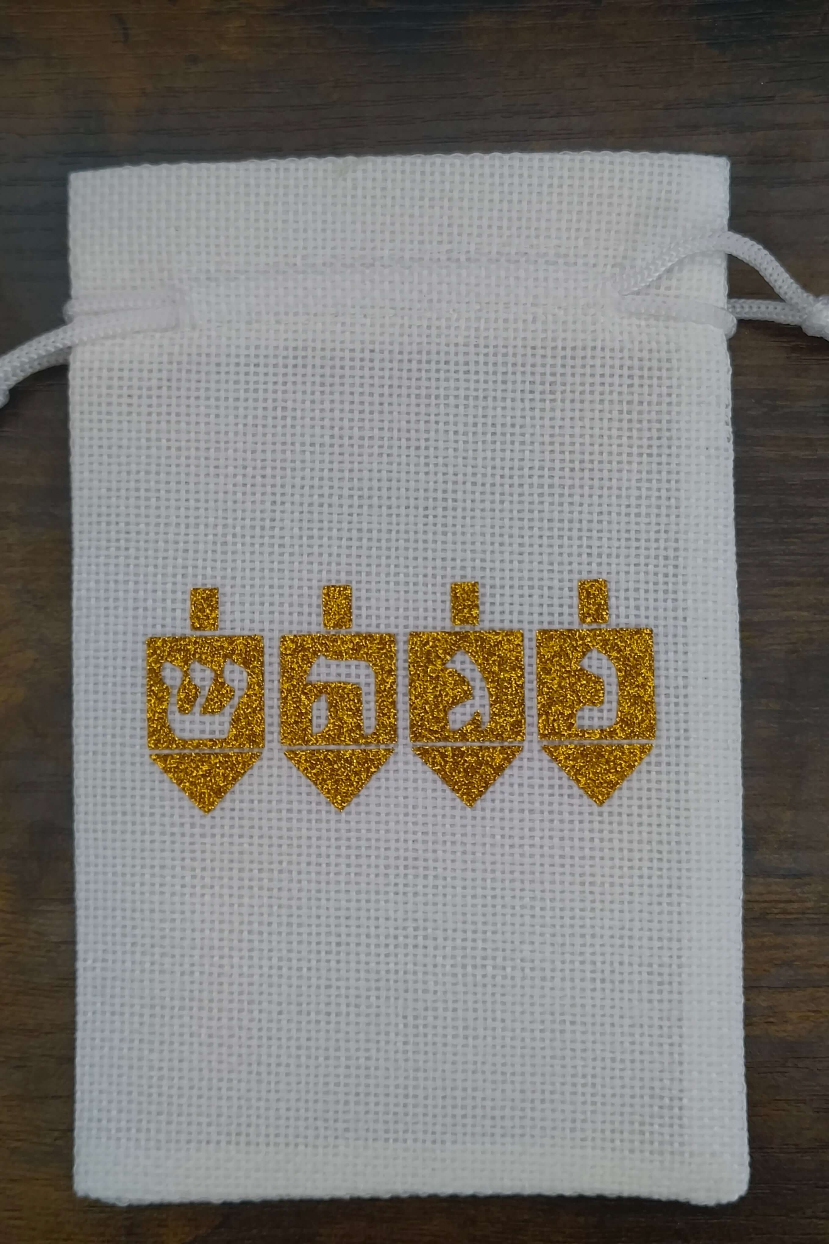 Handmade white drawstring hessian gift bag displayed on a dark wooden background. The front of the bag features four gold glitter dreidels, each with a Hebrew letter—Shin, Hey, Gimel, and Nun—representing the Hanukkah phrase “A great miracle happened there.” The festive design is cantered on the bag.