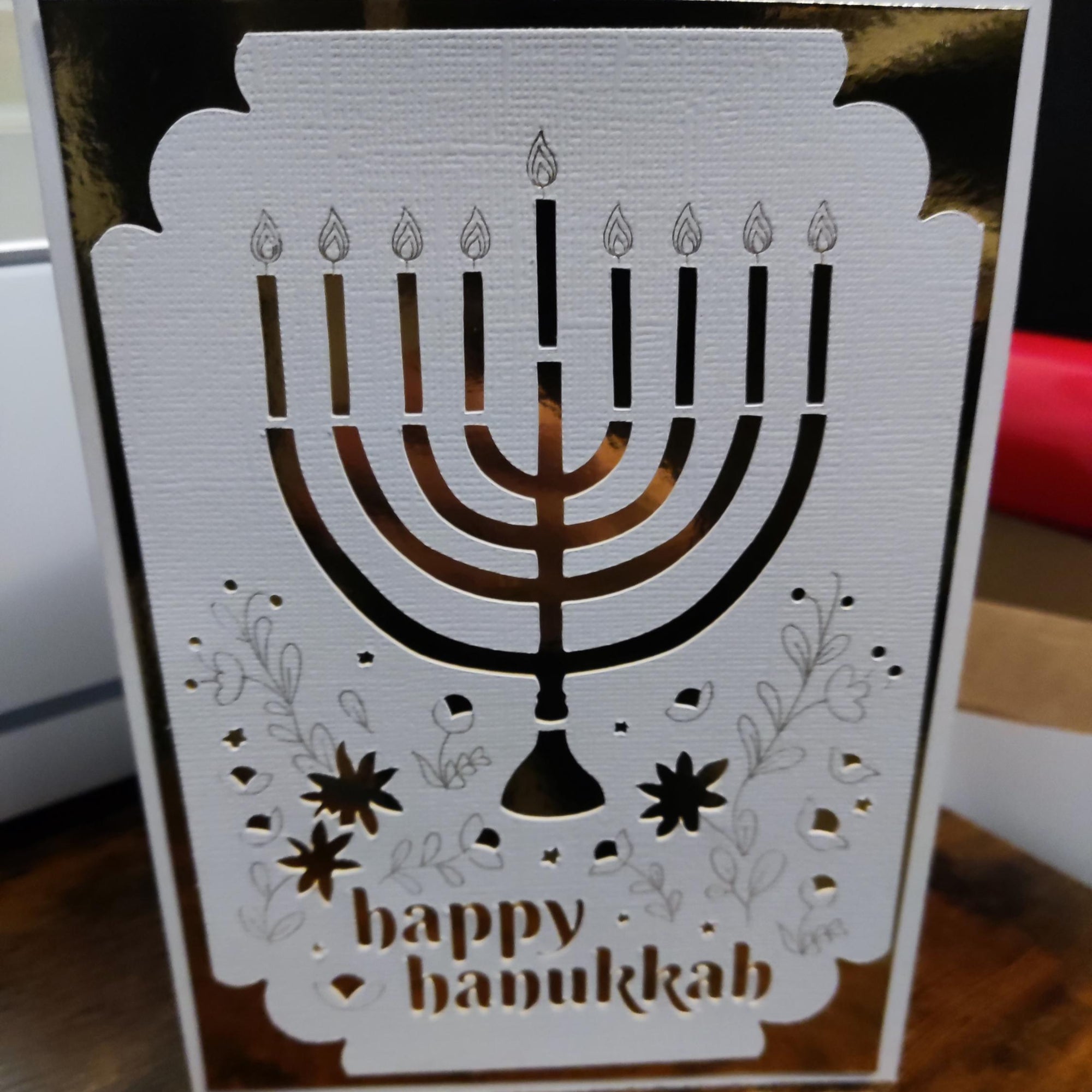 Elegant Hanukkah card with a gold menorah cut-out, floral decorations, and “Happy Hanukkah” text on textured white cardstock.