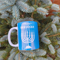 Happy Hanukkah mug with menorah design on blue background, surrounded by evergreen branches.