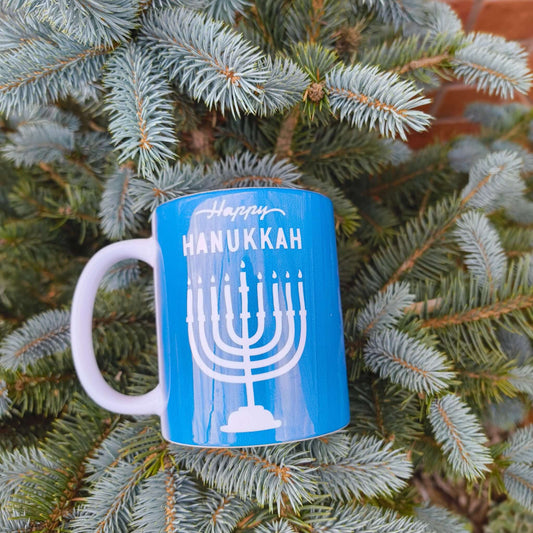 Happy Hanukkah mug with menorah design on blue background, surrounded by evergreen branches.