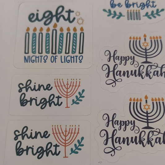 Close-up image of Hanukkah stickers featuring festive designs including “Eight Nights of Lights,” “Shine Bright” with menorahs, and “Happy Hanukkah” messages on a white sticker sheet.