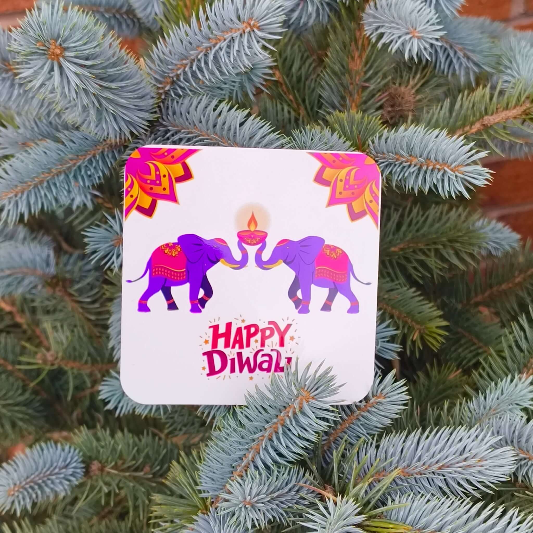 Close-up of a Happy Diwali coaster with two purple elephants and a glowing diya, resting on blue spruce branches.