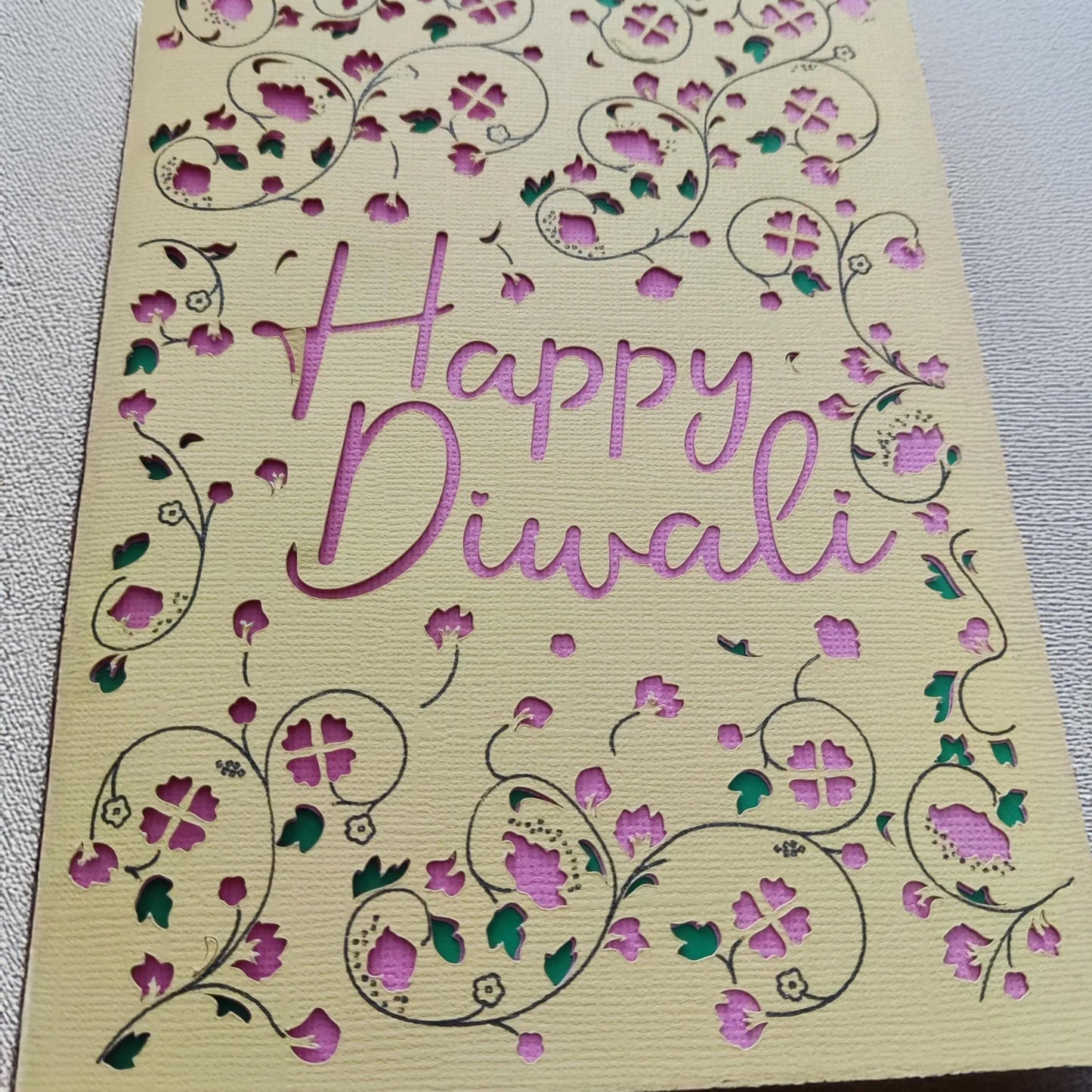 Close-up of a handmade Happy Diwali card featuring intricate floral cut-outs in pink and green against textured cream cardstock.