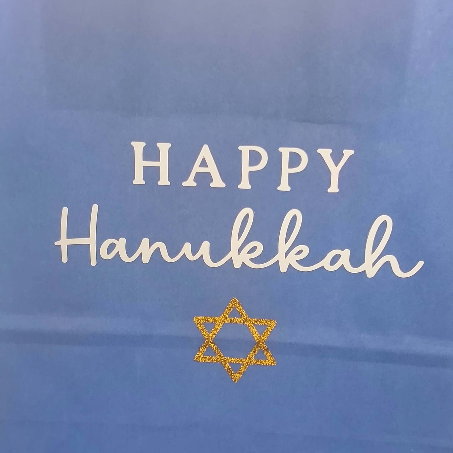Close-up of a blue Hanukkah gift bag with white 'Happy Hanukkah' text and a gold glitter Star of David.