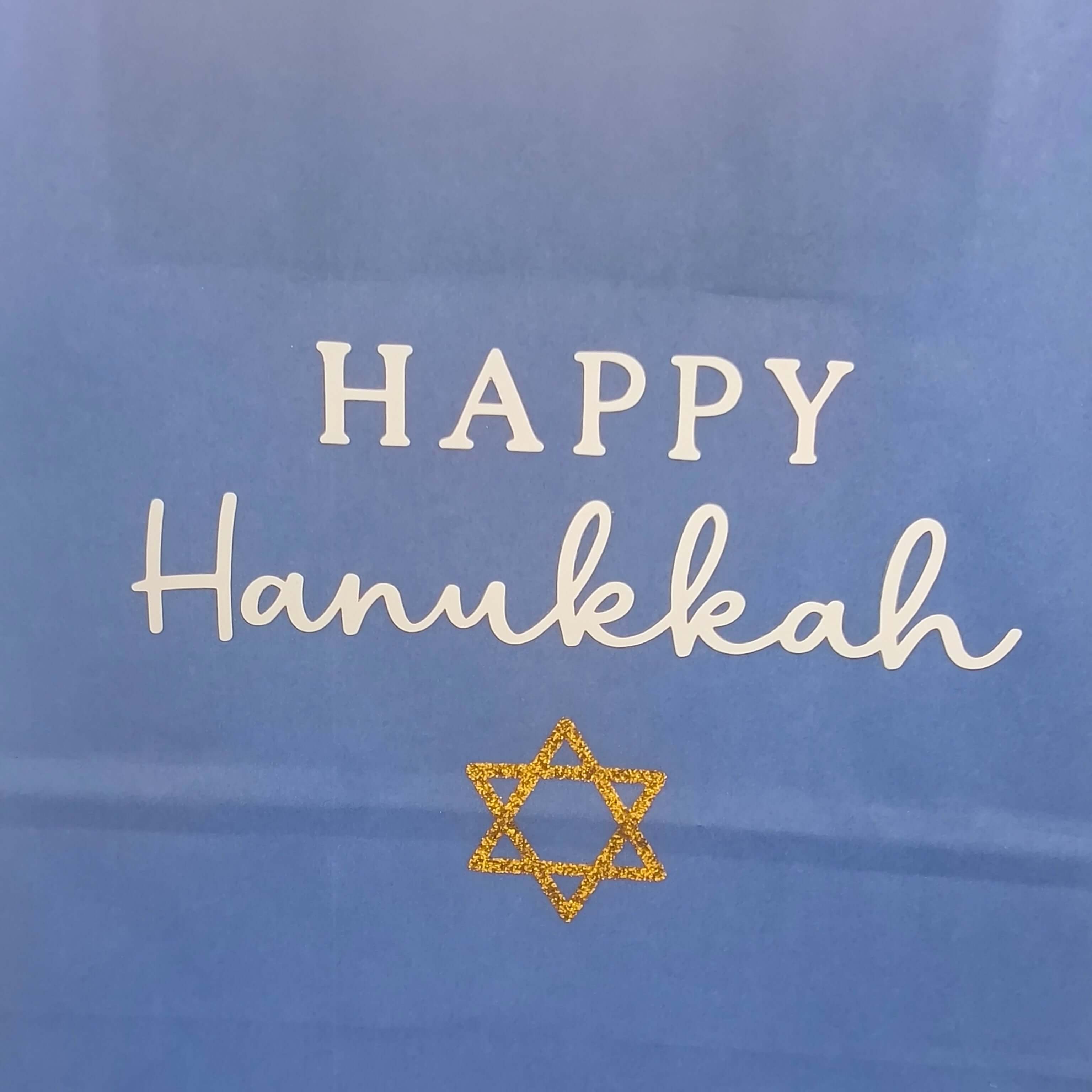 Close-up of a blue Hanukkah gift bag with white 'Happy Hanukkah' text and a gold glitter Star of David.