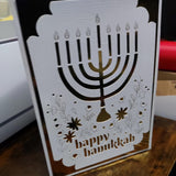 Festive Hanukkah greeting card with a gold foil menorah cut-out and delicate floral details, reading “Happy Hanukkah.”