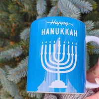 Happy Hanukkah mug with menorah design on blue ceramic coffee cup among evergreen branches.