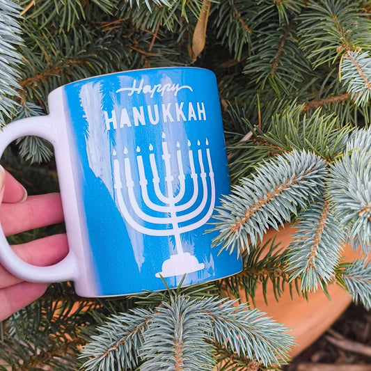 Happy Hanukkah mug displayed among green pine branches, featuring a blue design with a menorah.