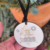Hand holding a ceramic “Happy New Year 2026” bauble with gold and black lettering and a black satin ribbon, displayed against evergreen foliage.