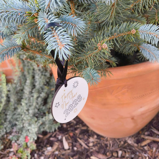 Round ceramic bauble reading “Happy New Year 2026” in gold and black text, hanging from a black satin ribbon on a blue spruce tree in a terracotta pot outdoors.