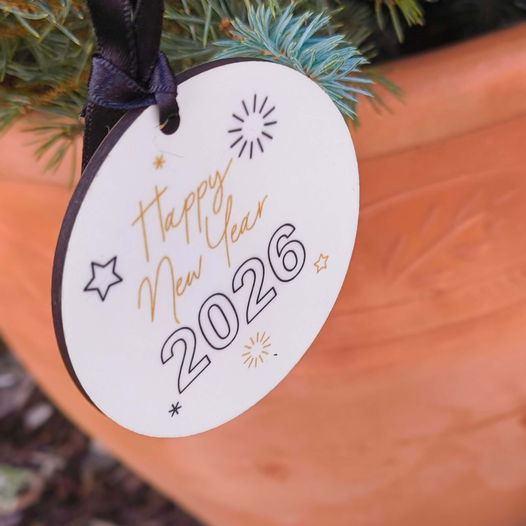 White ceramic “Happy New Year 2026” bauble with festive stars and fireworks design, hanging on a Christmas tree outdoors with a terracotta pot.