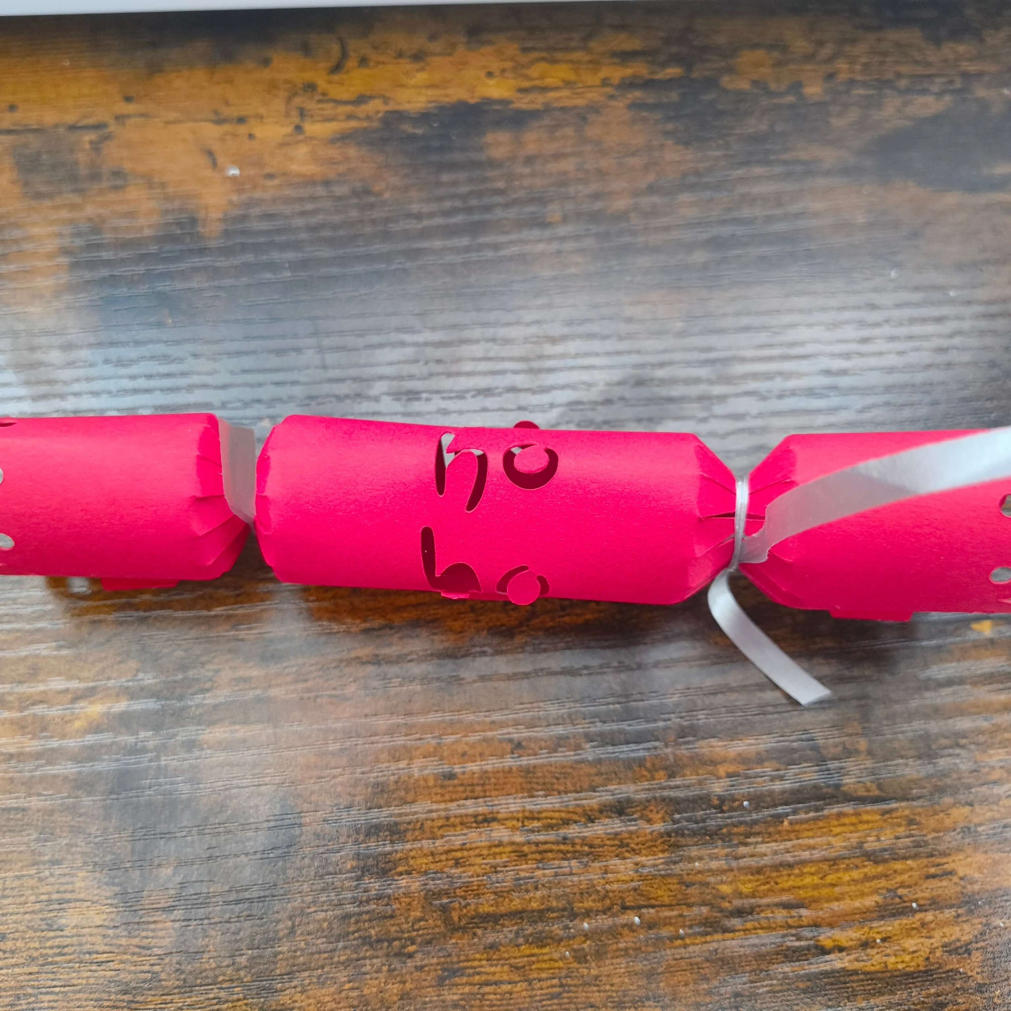Single red Christmas cracker with “Ho Ho” cut-out centre, silver ribbon ends, displayed on rustic wood background.