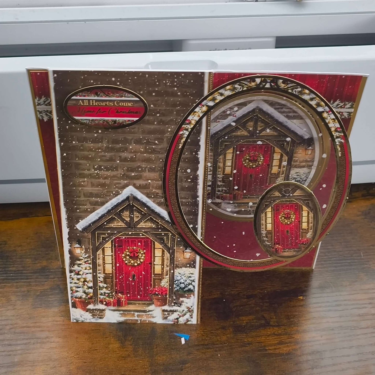 Handmade Christmas card featuring a cosy snow-covered cottage with a red front door and festive wreath, displayed on a wooden table.