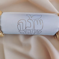 Close-up of a white Rosh Hashanah cracker with gold foil Hebrew lettering and apple motif, set against soft fabric.
