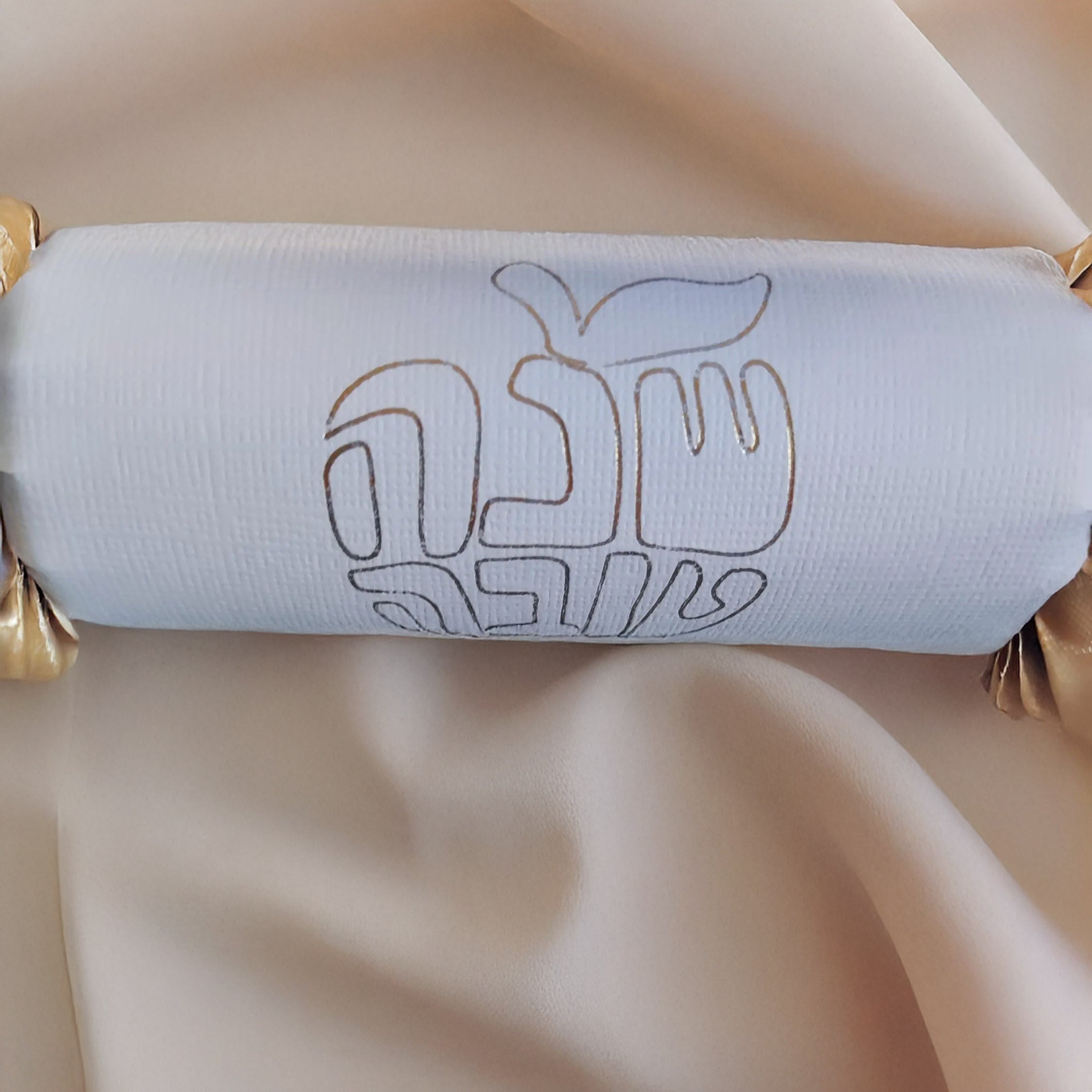 Close-up of a white Rosh Hashanah cracker with gold foil Hebrew lettering and apple motif, set against soft fabric.
