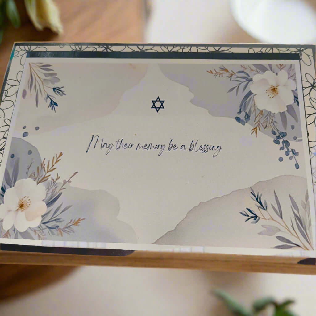 Jewish sympathy card with Star of David and floral border, text reads 'May their memory be a blessing', soft pastel design on wooden surface