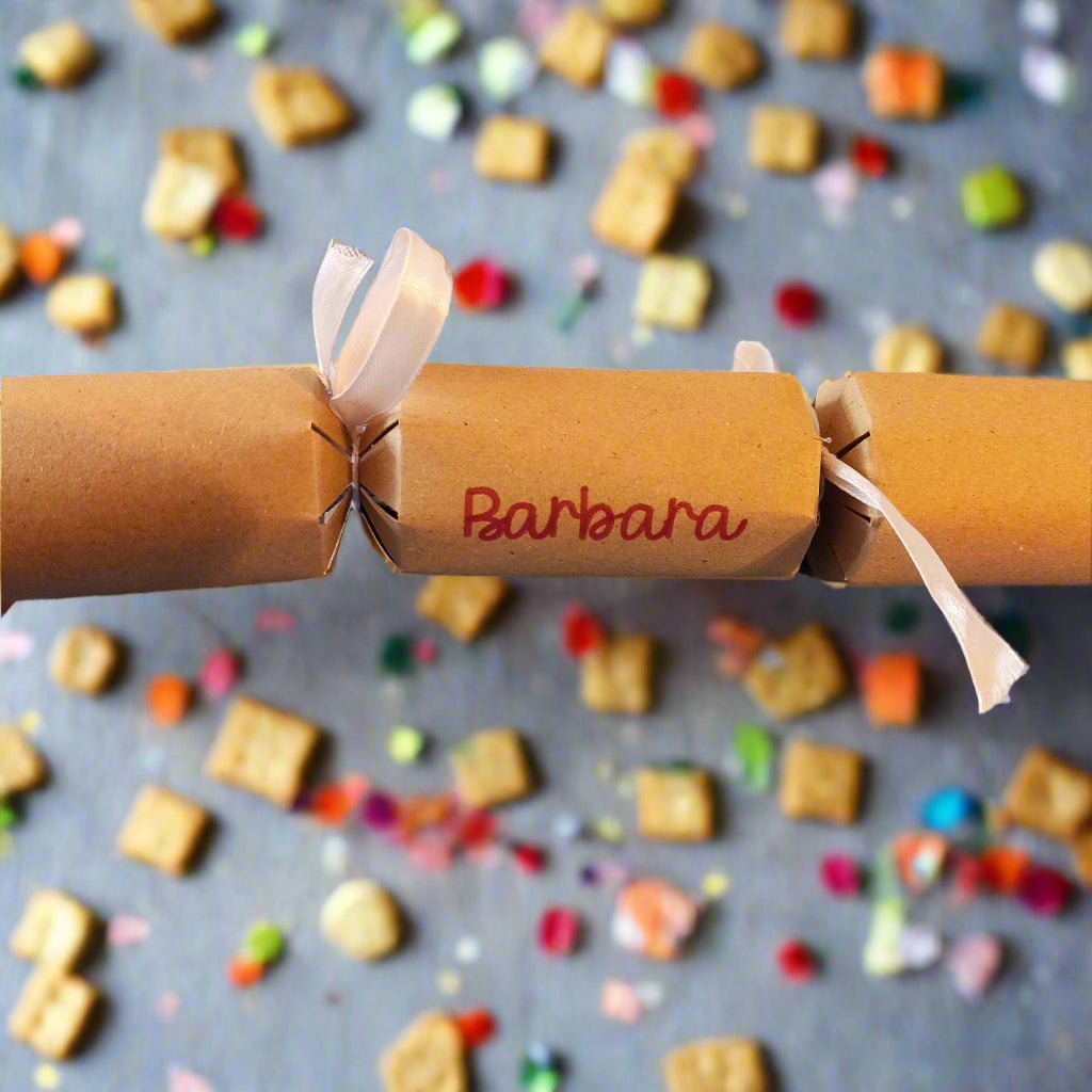 Personalised kraft cracker with “Barbara” in red lettering, tied with white ribbon, photographed against a playful backdrop of colourful sweets and biscuits.