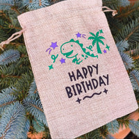 Large hessian Happy Birthday bag with dinosaur, palm tree, and stars design, photographed on blue spruce tree.
