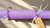 Handmade personalised Diwali cracker in lavender with white ribbon and gold tag reading 'Diwali Blessings', featuring custom name space in silver foil on a sunlit surface.
