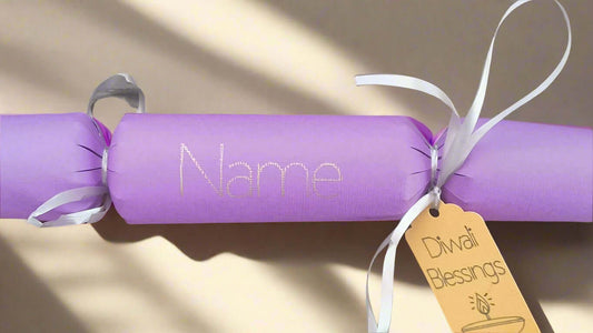 Handmade personalised Diwali cracker in lavender with white ribbon and gold tag reading 'Diwali Blessings', featuring custom name space in silver foil on a sunlit surface.