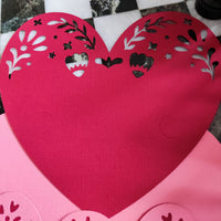 Close-up of layered heart papercut in red and pink textured cardstock, partially assembled, viewed from above on black and white tile background, handmade romantic craft