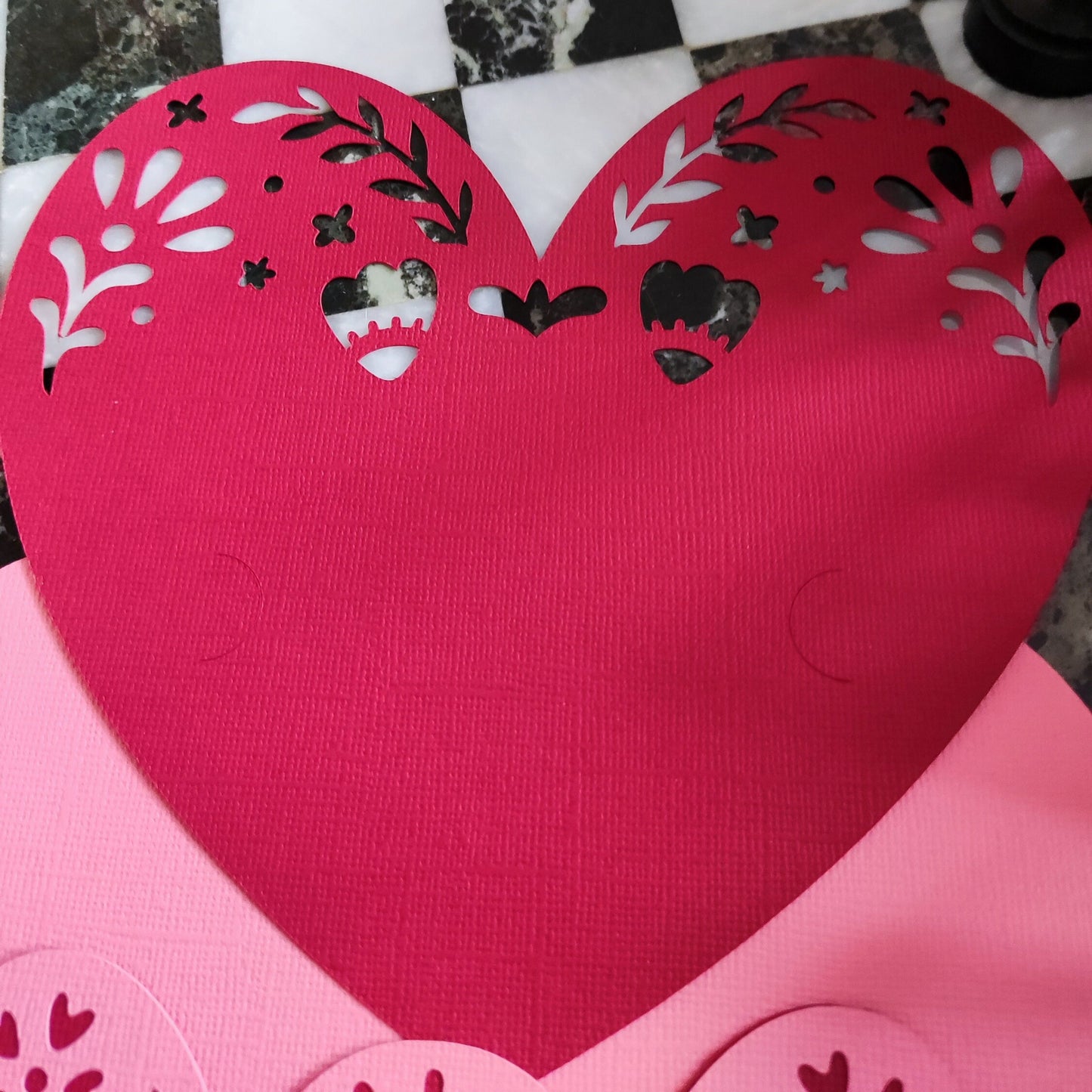 Close-up of layered heart papercut in red and pink textured cardstock, partially assembled, viewed from above on black and white tile background, handmade romantic craft