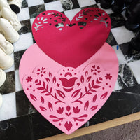 Layered heart papercut in progress, pink and red textured cardstock with floral cutouts, top-down angle on black and white chessboard surface, handmade romantic craft