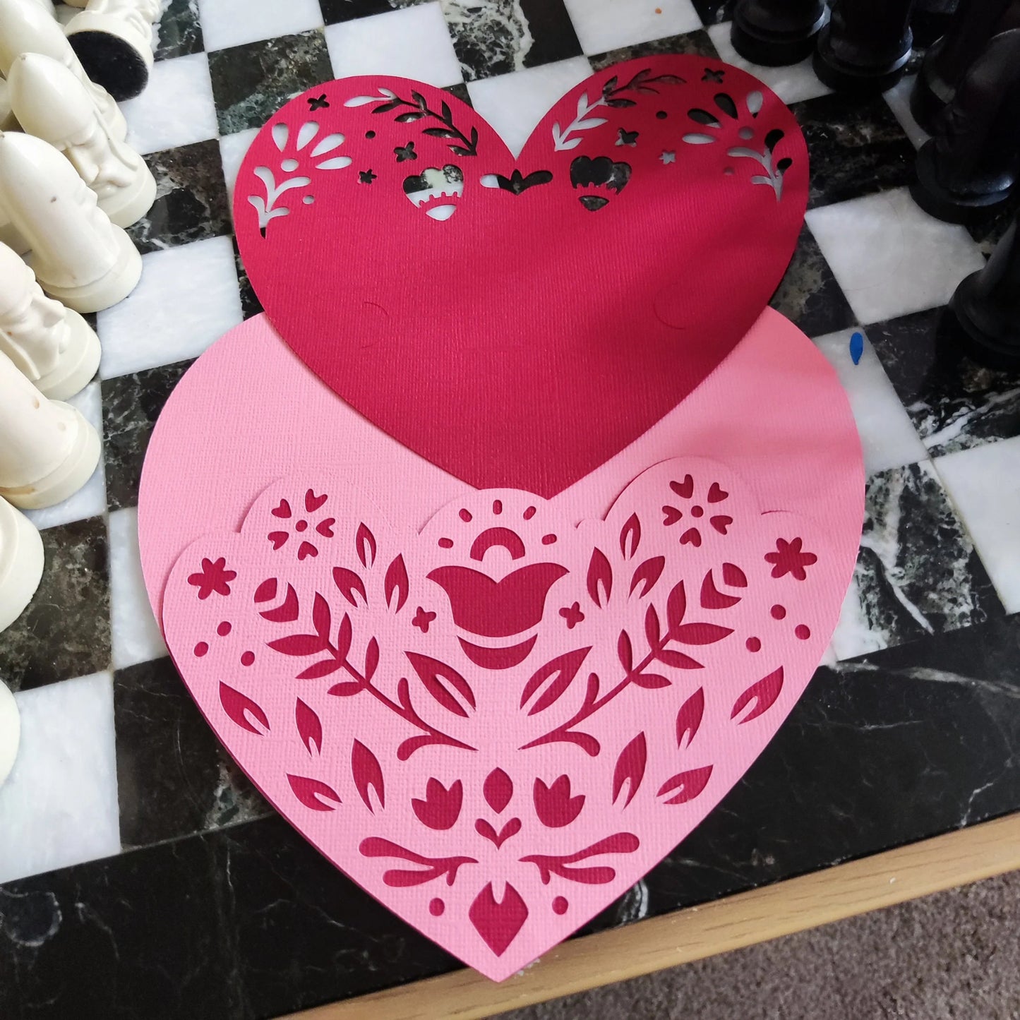 Layered heart papercut in progress, pink and red textured cardstock with floral cutouts, top-down angle on black and white chessboard surface, handmade romantic craft