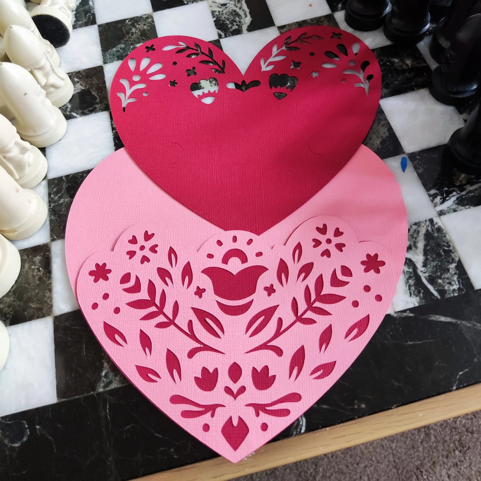 Layered heart papercut in progress, pink and red textured cardstock with floral cutouts, top-down angle on black and white chessboard surface, handmade romantic craft