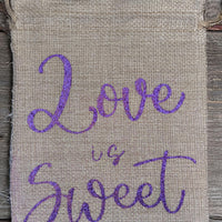 Small rustic hessian drawstring bag with “Love is Sweet” in purple glitter script. Shown with fabric hearts for scale. Natural woven texture adds a handmade charm.