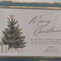 Elegant handmade Christmas card with a dark green textured border, gold foil trim, and a central image of a Christmas tree topped with a gold star and surrounded by white gift boxes. The card features the message “Merry Christmas” in flowing script and a heartfelt blessing message below, signed “Love the Baxter Family.” Displayed flat on a wooden surface.

