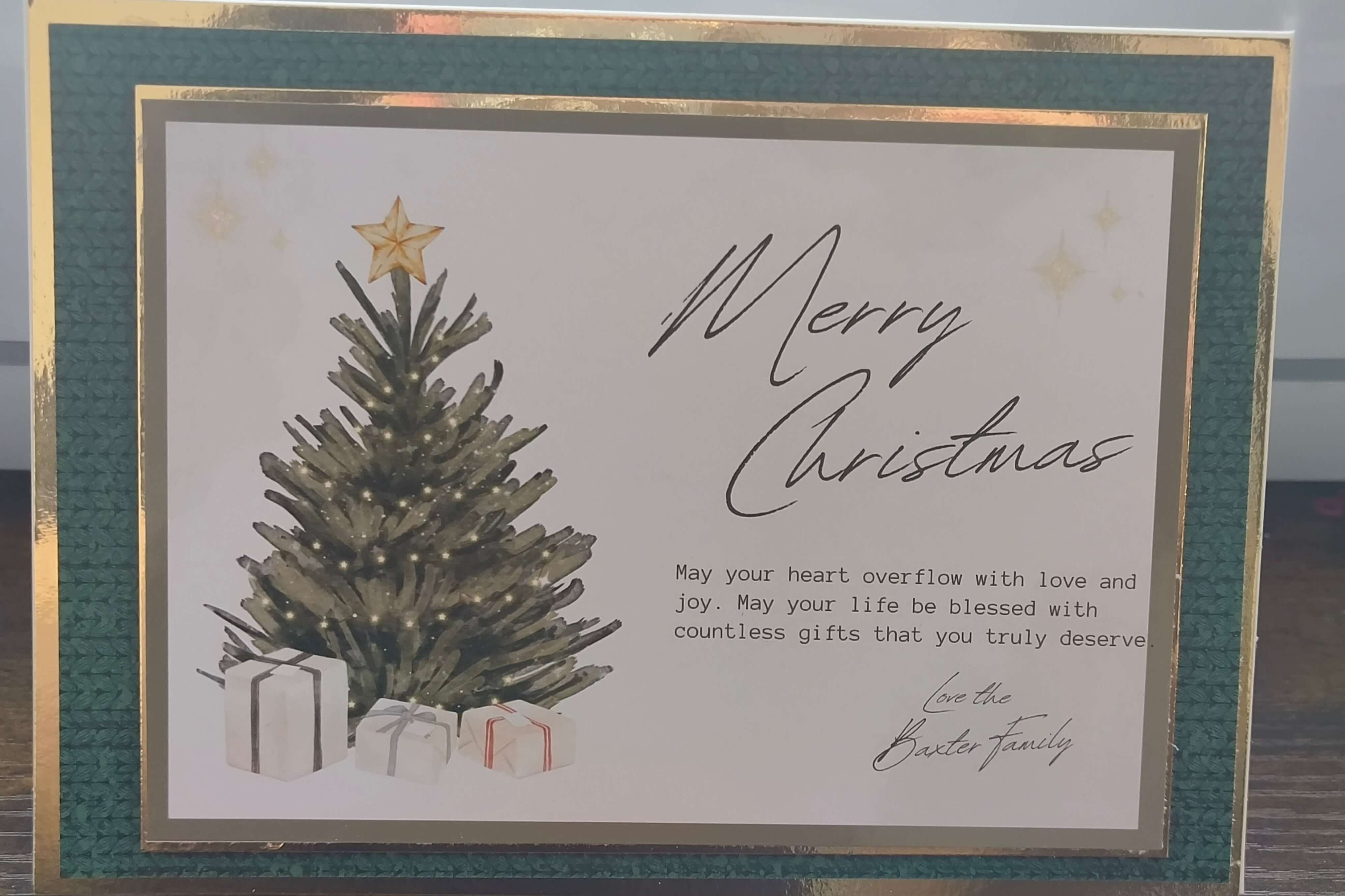 Elegant handmade Christmas card with a dark green textured border, gold foil trim, and a central image of a Christmas tree topped with a gold star and surrounded by white gift boxes. The card features the message “Merry Christmas” in flowing script and a heartfelt blessing message below, signed “Love the Baxter Family.” Displayed flat on a wooden surface.

