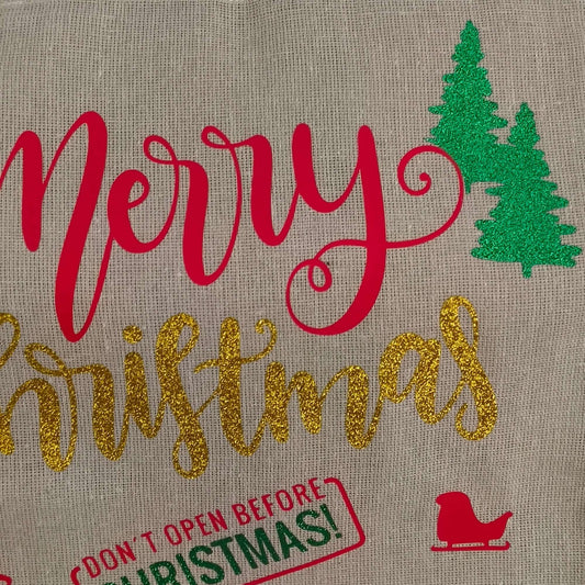 Close-up of glitter Merry Christmas design on hessian sack – red “Merry”, gold glitter “Christmas”, green vinyl trees, and festive sleigh icon, perfect for personalised holiday gift bags.