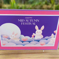 Angled outdoor photo of a handmade Mid Autumn Festival card featuring moon rabbits and lotus flowers, placed on a wooden table with a garden background.
