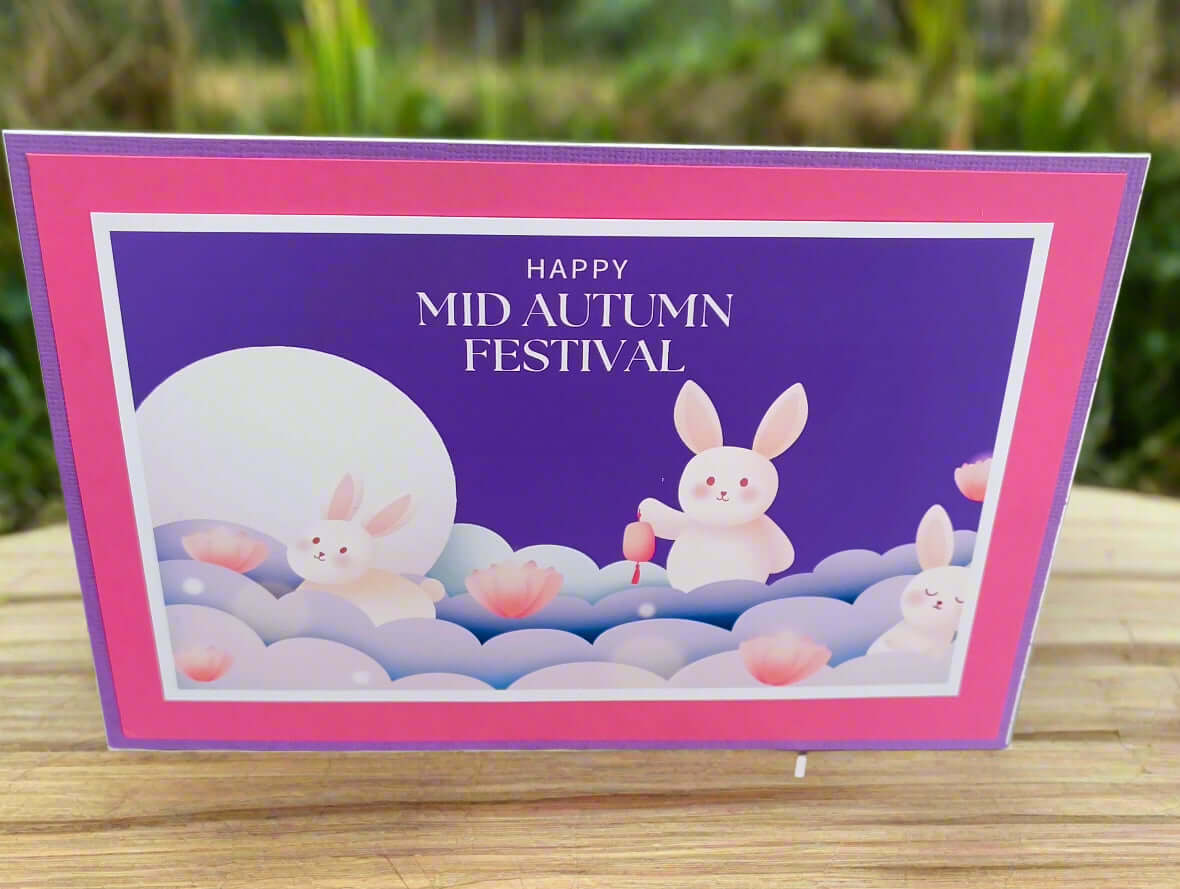 Angled outdoor photo of a handmade Mid Autumn Festival card featuring moon rabbits and lotus flowers, placed on a wooden table with a garden background.