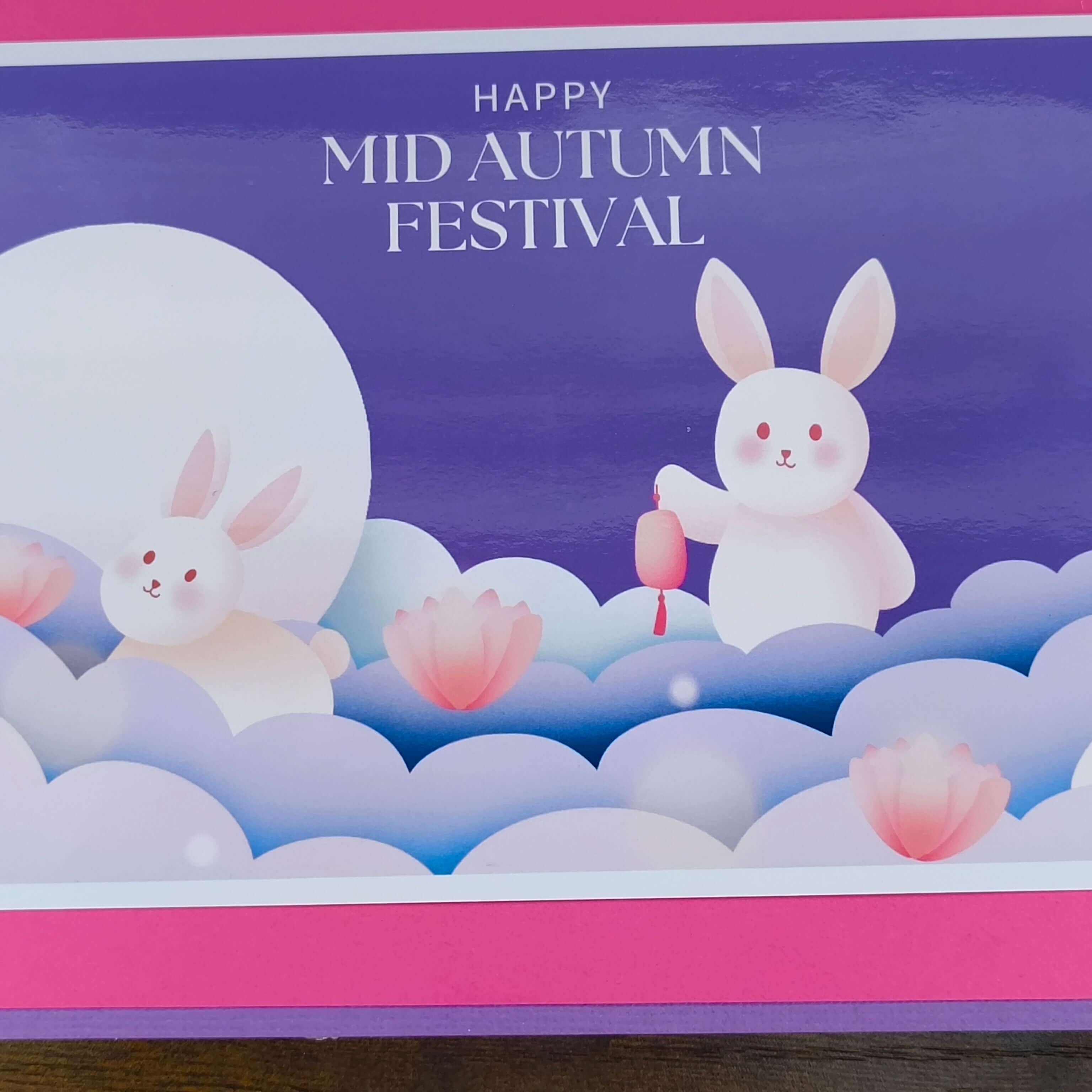 Close-up of a handmade Mid Autumn Festival card featuring pastel moon rabbits, pink lotus flowers, and a glowing full moon on a purple background.