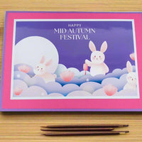 Flat lay top-down view of a handmade Mid Autumn Festival card with cute moon rabbits and lotus flowers, styled on a bamboo mat with red lanterns, chopsticks, and decorative elements.