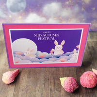 Front-facing angle of a handmade Mid Autumn Festival card featuring cute moon rabbits, lotus flowers, and a dreamy moonlit background, styled on a wooden table with decorative lychee pods.