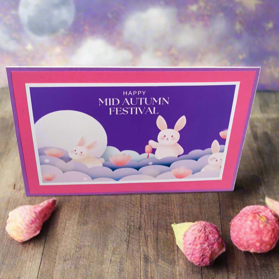 Front-facing angle of a handmade Mid Autumn Festival card featuring cute moon rabbits, lotus flowers, and a dreamy moonlit background, styled on a wooden table with decorative lychee pods.