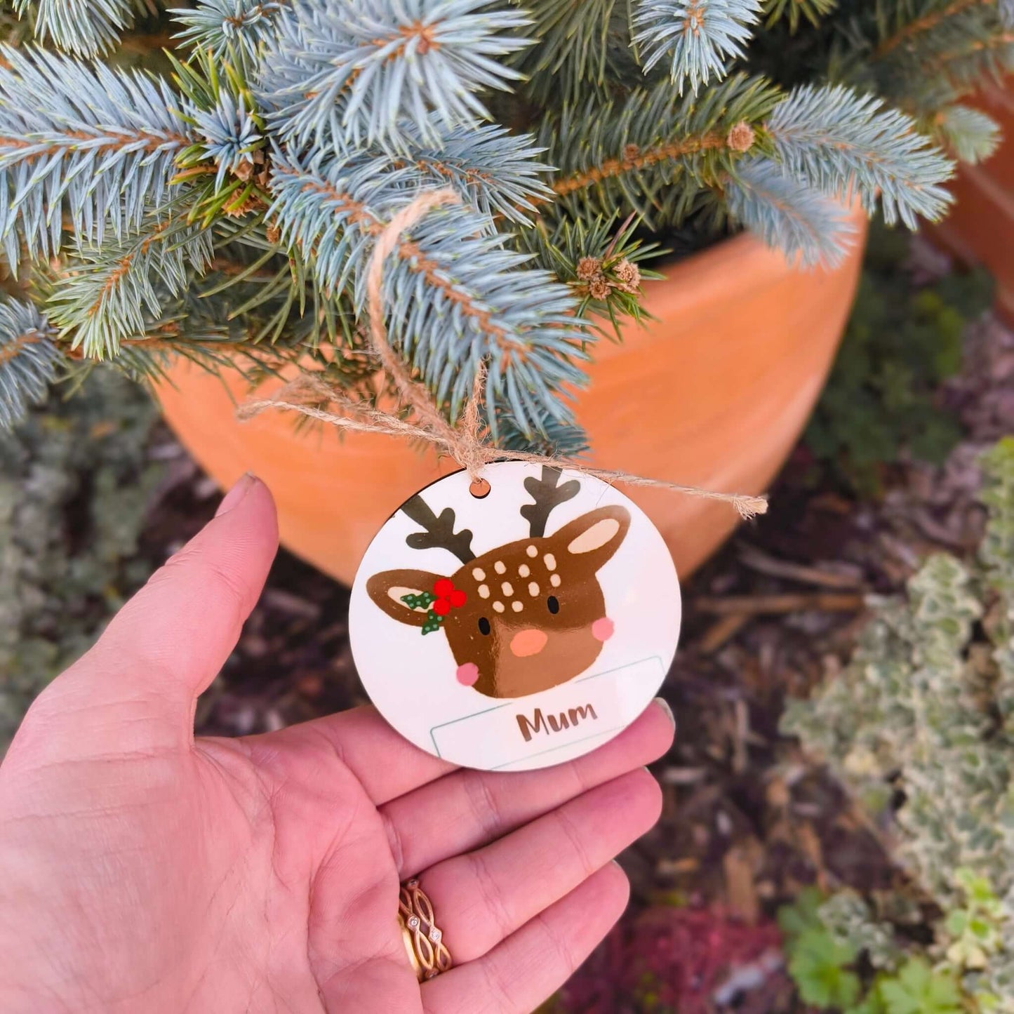 Personalised reindeer bauble with ‘Mum’ text held in hand against outdoor pine tree background
